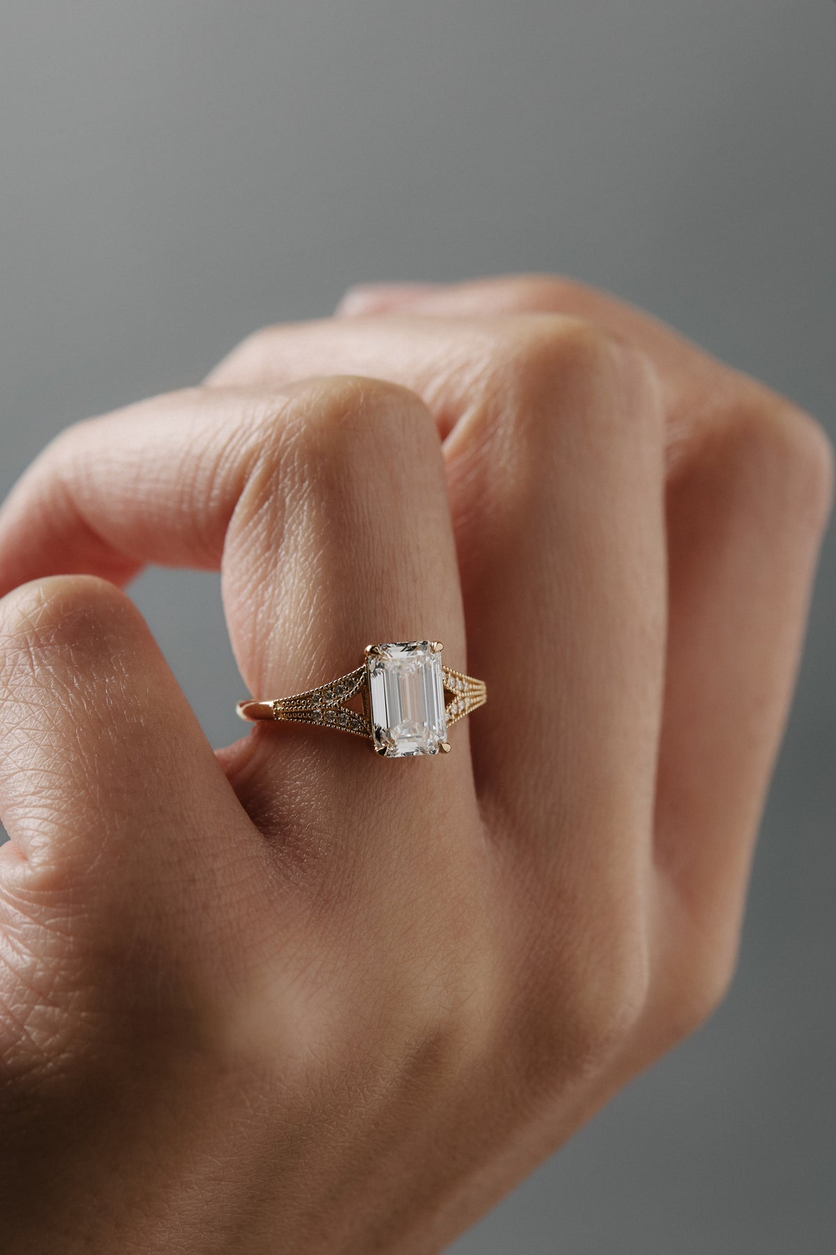 Close-up of the Cora split shank milgrain engagement solitaire worn on a woman's finger, featuring an emerald cit diamond and round diamond accents.