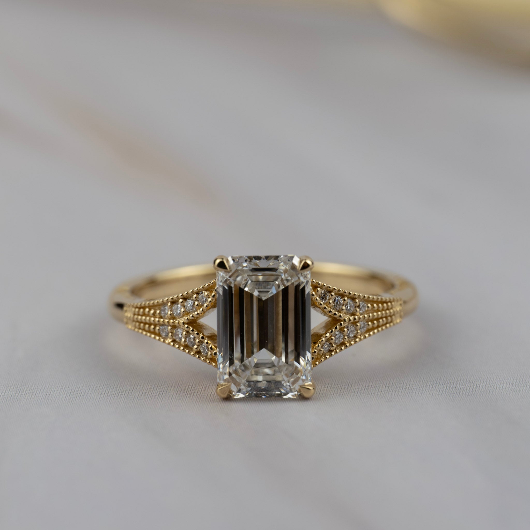 Cora split-shank engagement solitaire featuring an emerald cut diamond accented by round diamonds in a detailed milgrained gold band.