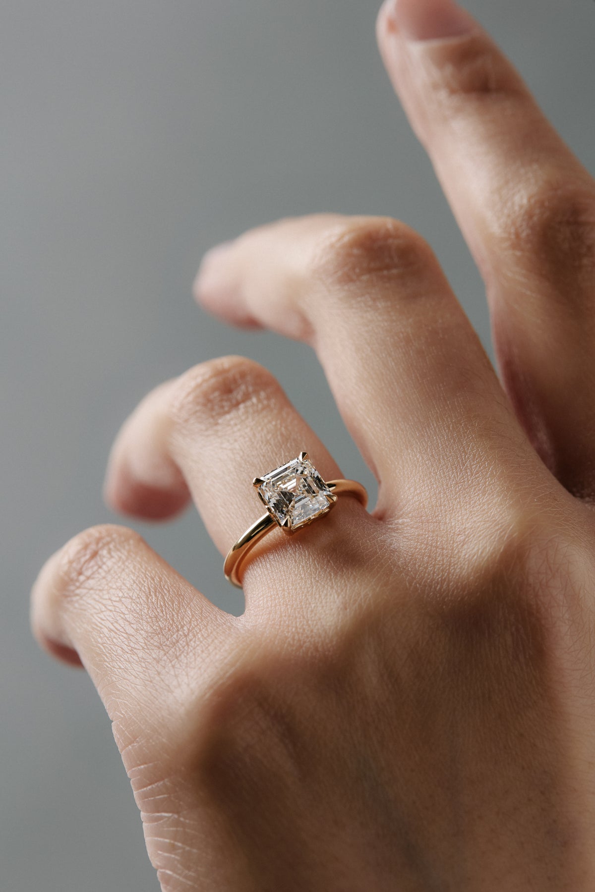Asschercut diamond claw solitaire engagement ring with floral scroll side detailing, worn alone on a woman's finger.
