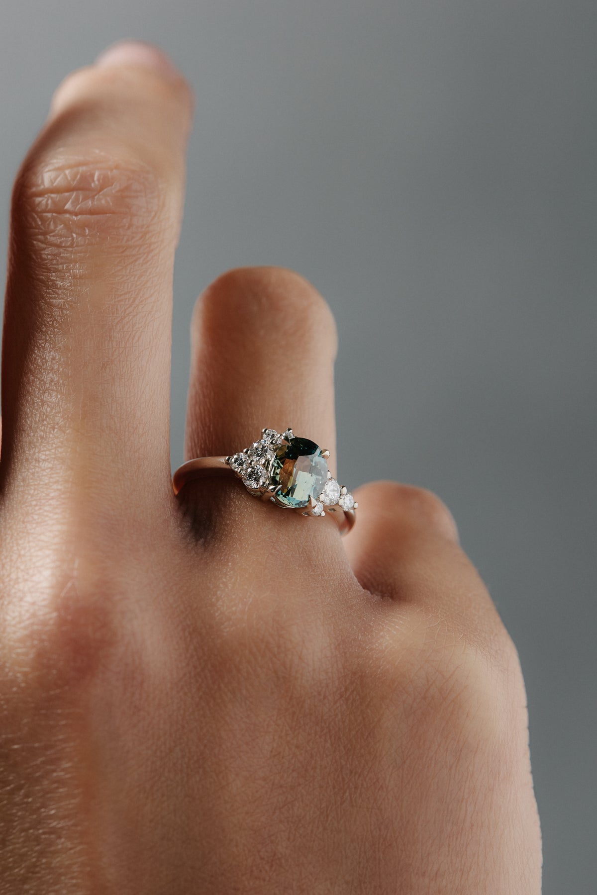 Close-up of the Celeste teal sapphire and Canadian diamond cluster engagement ring worn a woman's finger. 