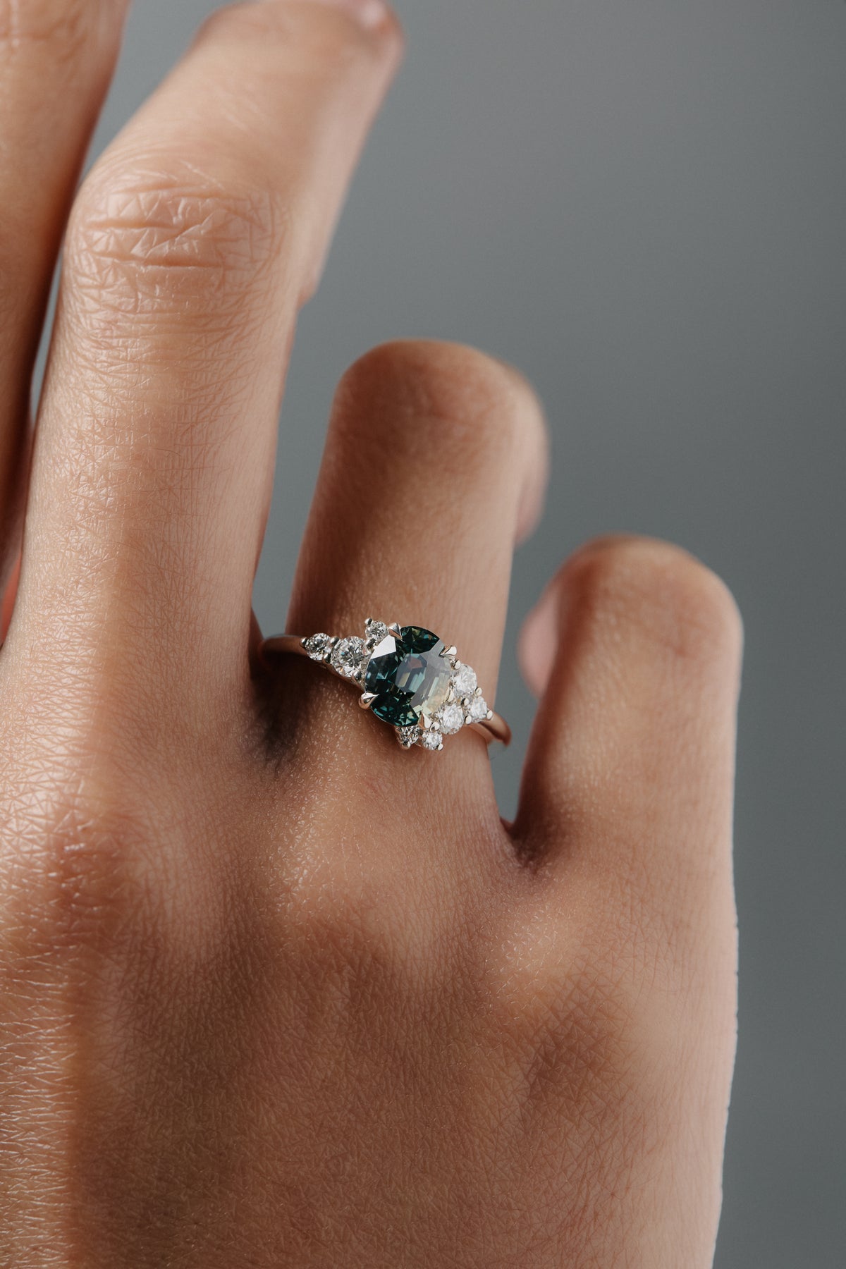 Close-up of a woman wearing the Celeste teal sapphire and diamond cluster engagement ring on her finger. 