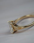 Close-up side profile o father Cora split-shank milgrain engagement solitaire showing the emerald cut diamond, round diamond accents, and detailed gold setting.