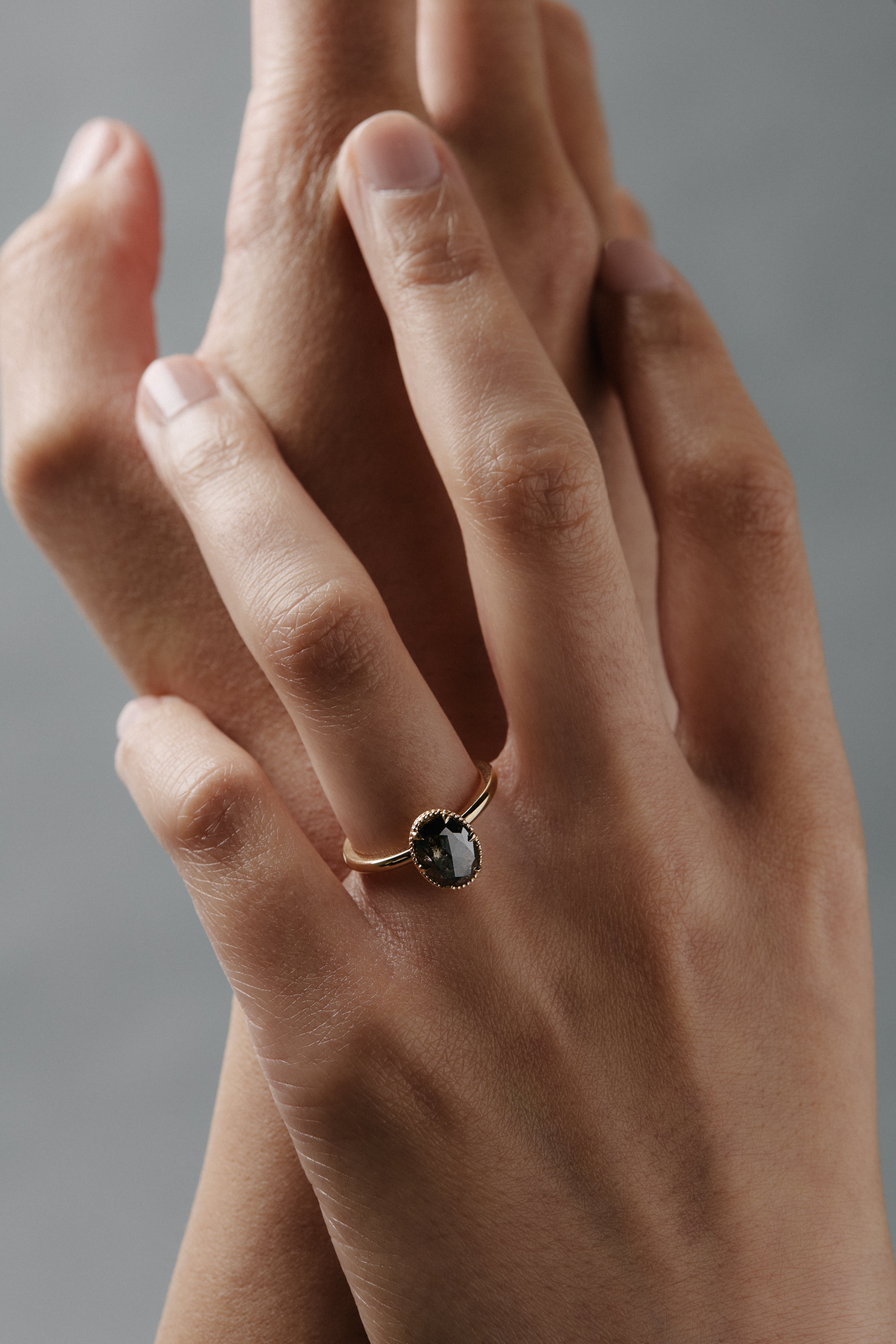 Stylized close-up of women's hands wearing the Elanor salt and pepper oval diamond six-claw solitaire with an Art Deco woven basket and beaded edge.
