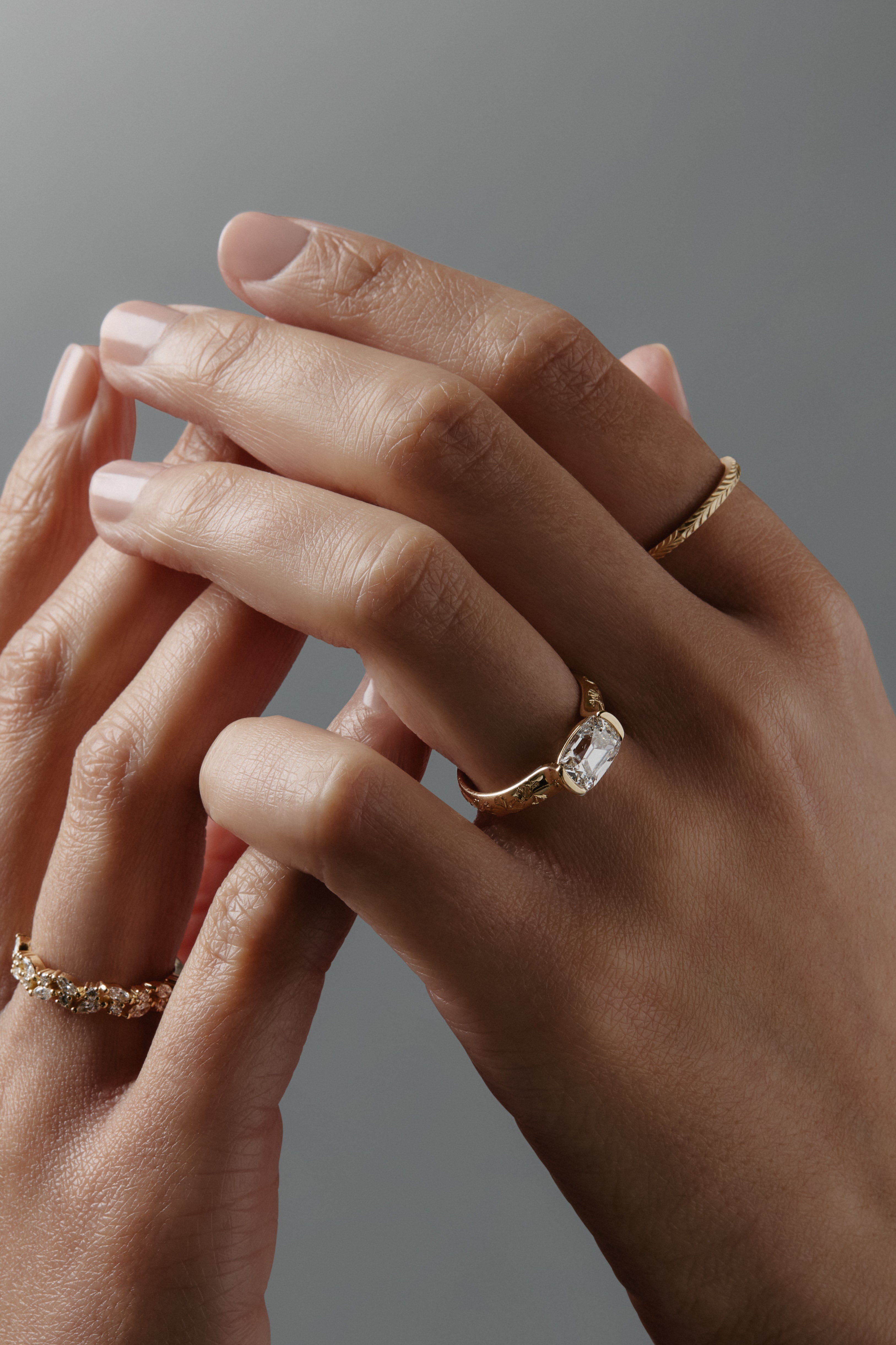 Close-up of hands wearing multiple gold rings, including a thin Fairmined yellow gold wedding band with hand-engraved fern details.