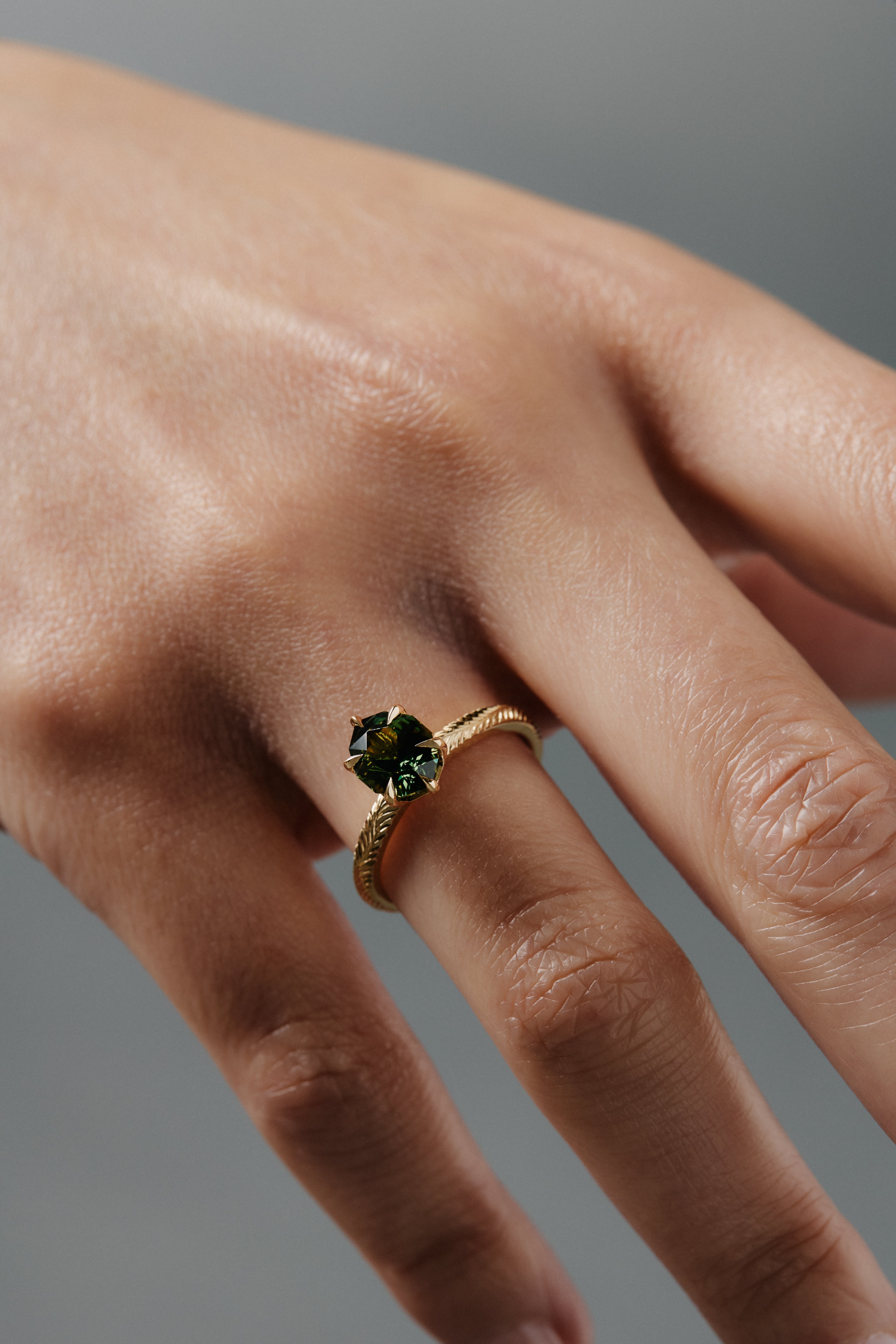 Close-up of a woman wearing the Fern hand engraved solitaire in Fairmined gold, featuring a step cut oval green sapphire.