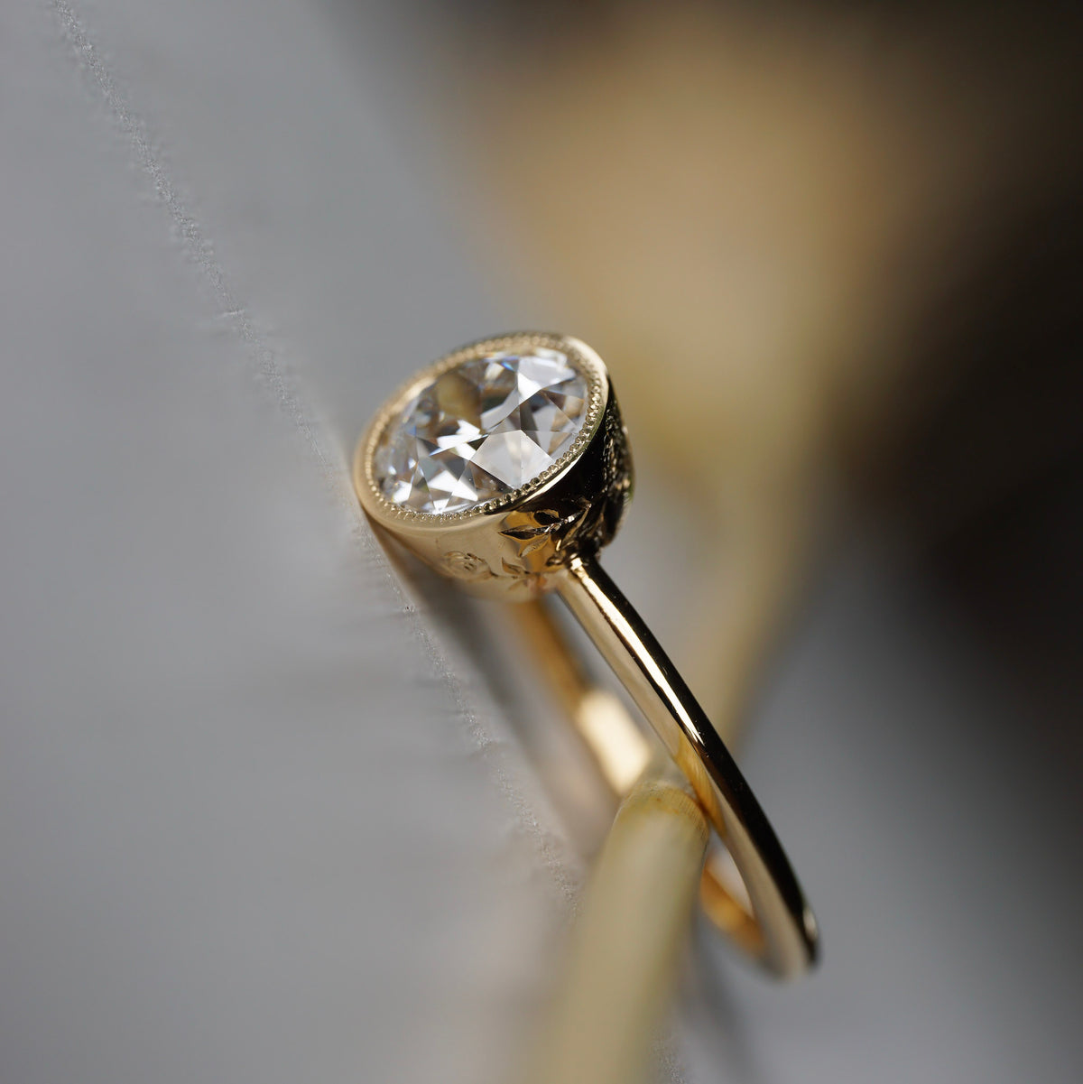 Flora gold milgrain bezel solitaire featuring an Old European cut diamond with floral engravings along the sides.