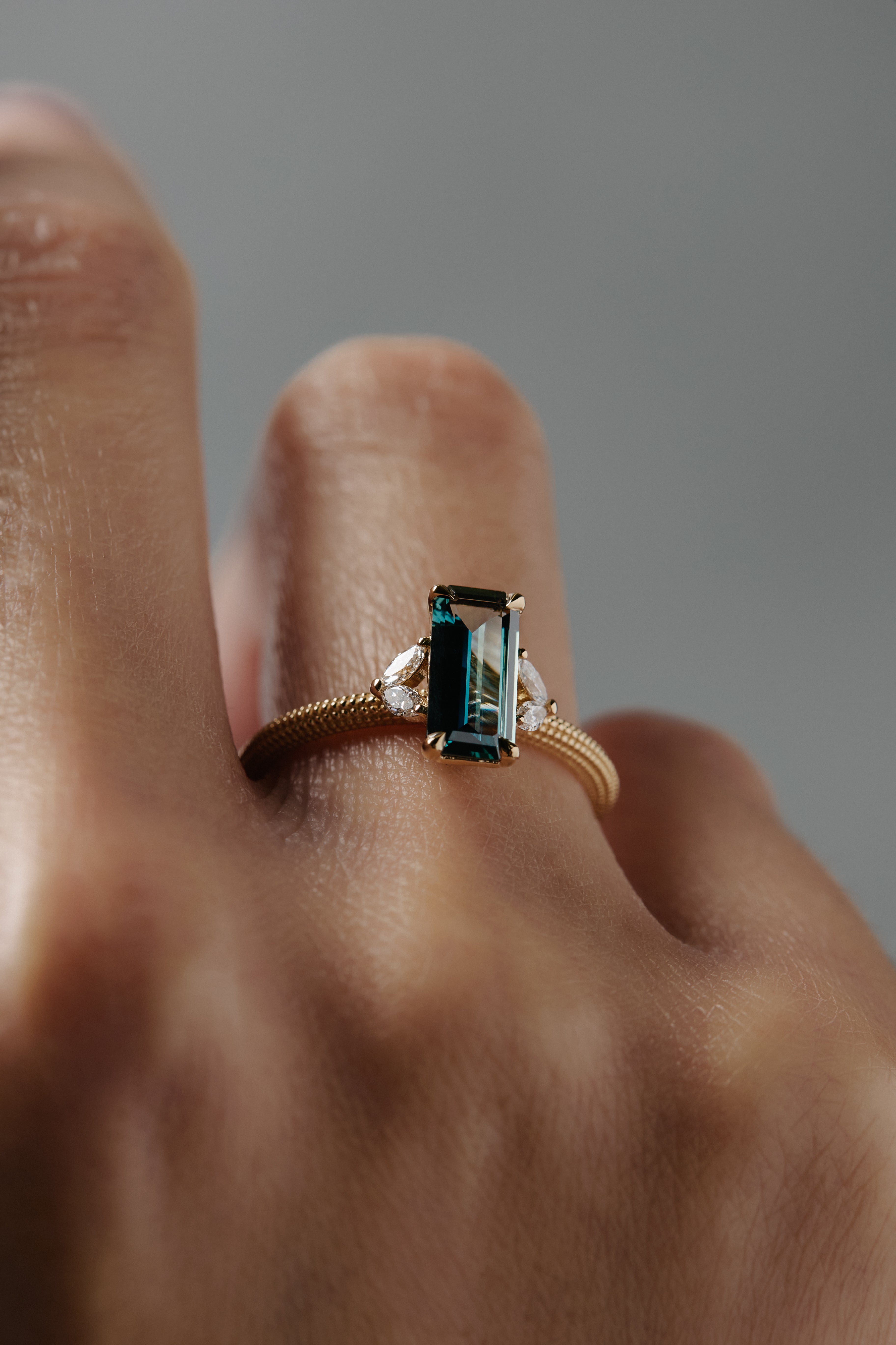 Close-up of a woman wearing the Millie cluster solitaire, highlighting the rectangular teal sapphire, marquise diamonds- and intricate milgrained beaded band 