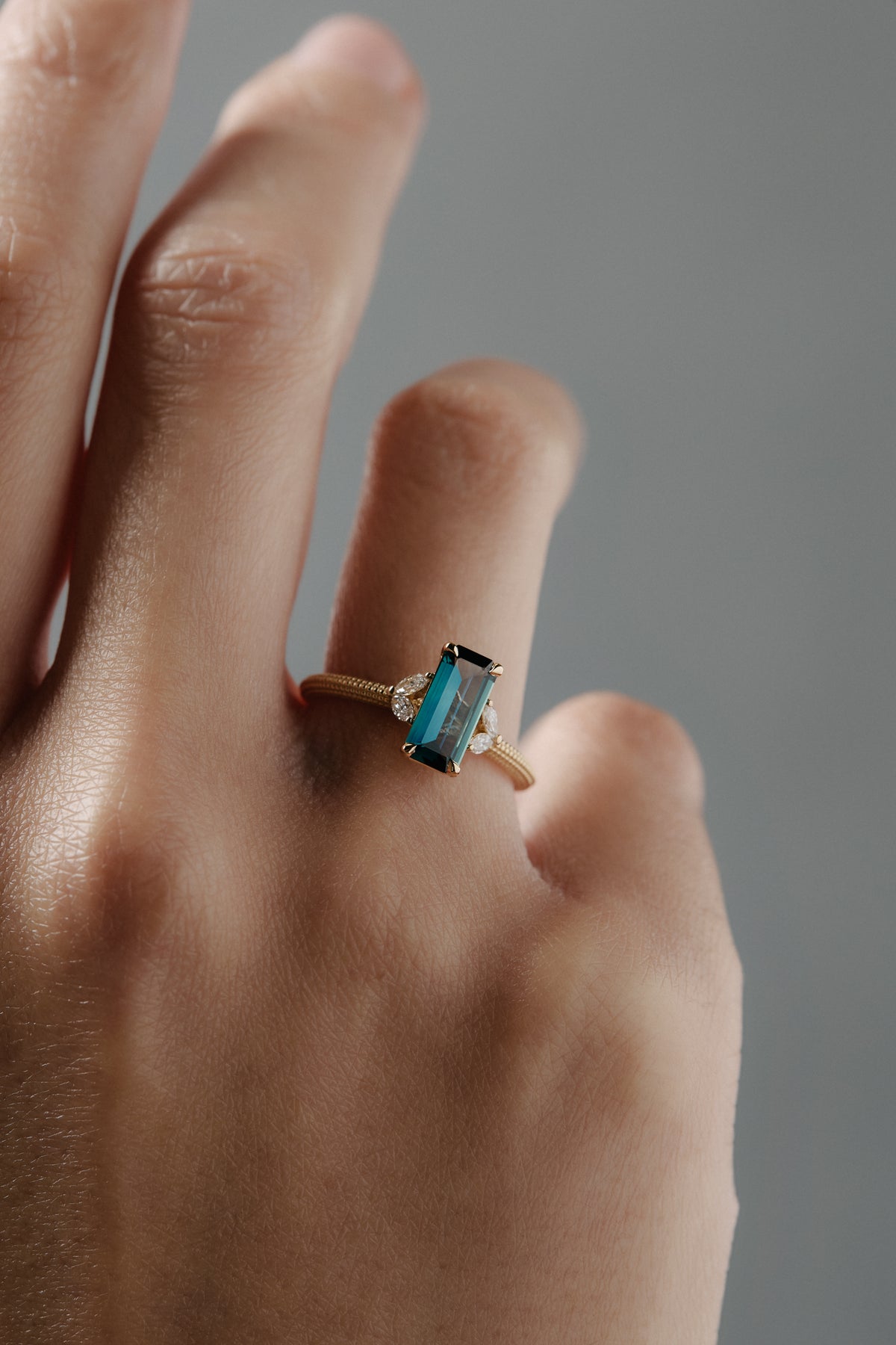Close-up of the Millie cluster solitaire engagement ring worn on a woman's finger, featuring a rectangular teal sapphire, marquise diamonds, and a milgrained beaded band.