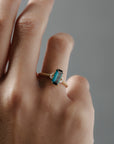 Close-up of the Millie cluster solitaire engagement ring worn on a woman's finger, featuring a rectangular teal sapphire, marquise diamonds, and a milgrained beaded band.