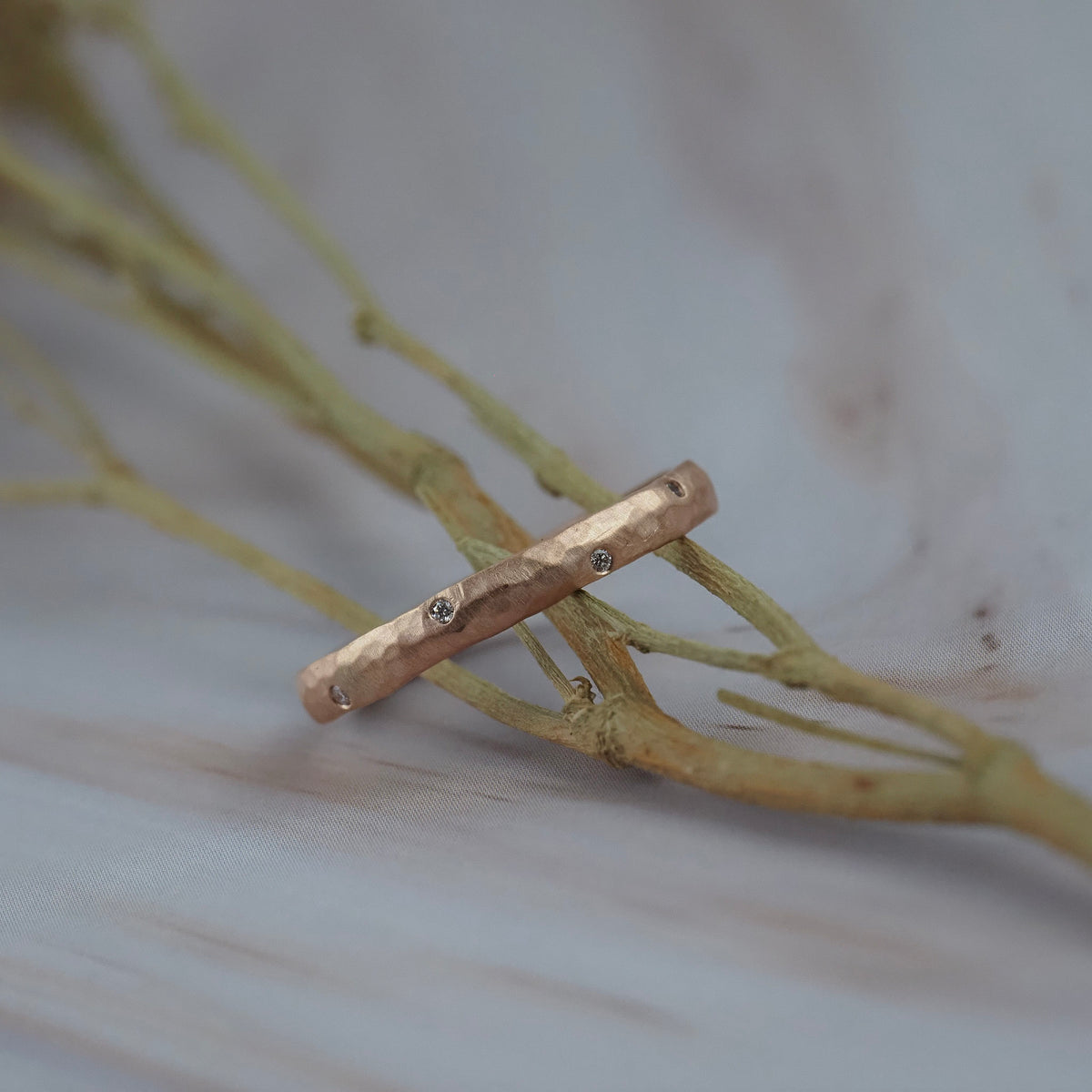 Starlight  hammered rose gold wedding band ring with small round diamonds, resting on dried beige branches over a light gray marble surface.